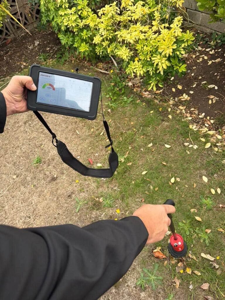Technician holding a handheld leak detector with a screen and a ground microphone to pinpoint an underground water leak in a garden